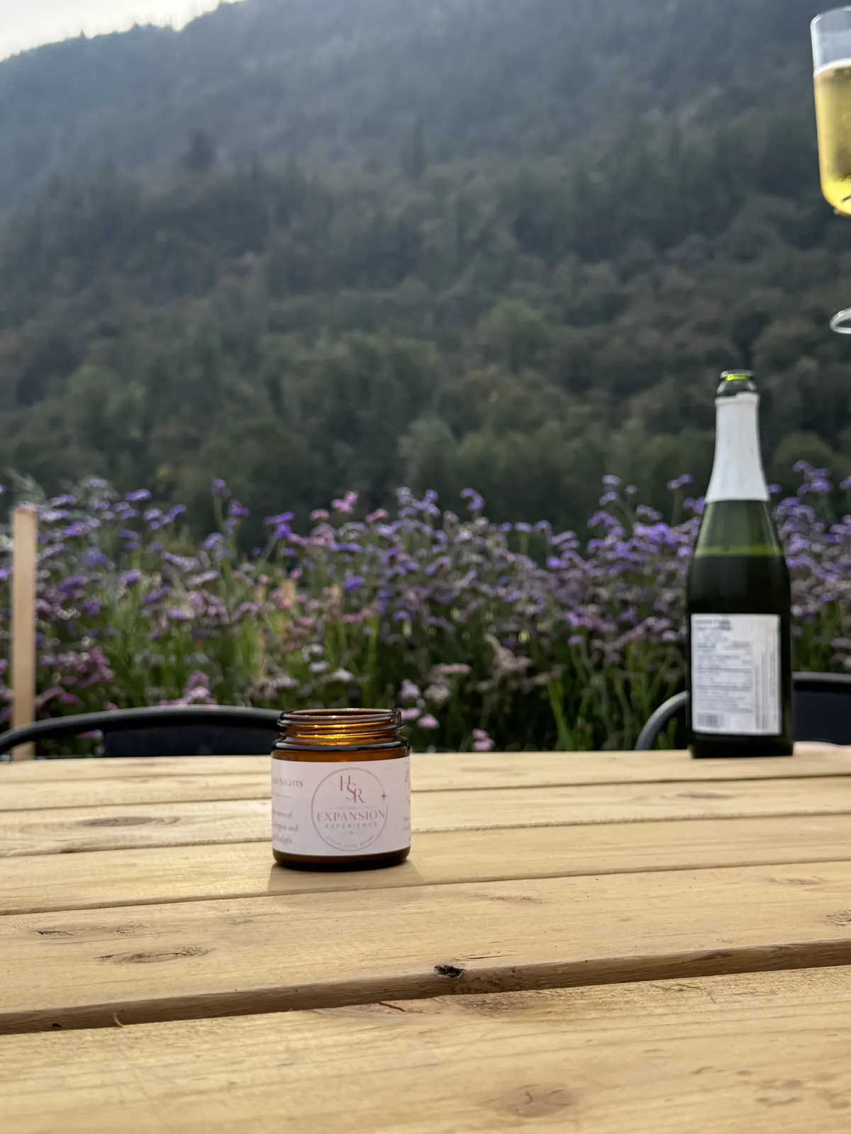 A lit candle with a blush custom label on a wooden table, with purple wildflowers and a forested mountain behind